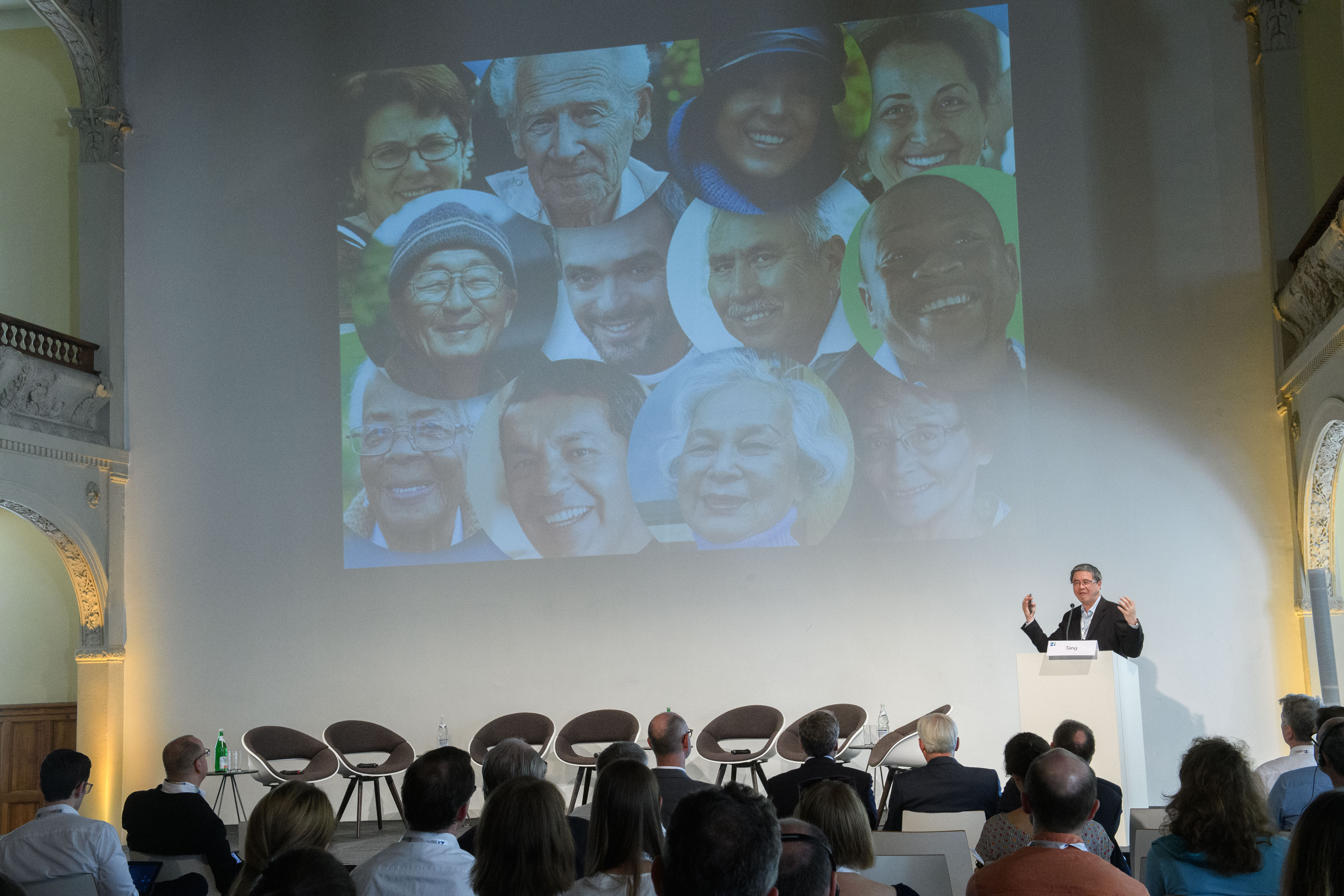 Herr Tang hält einen Vortrag vor Publikum, es werden Gesichter an die Wand projiziert.
