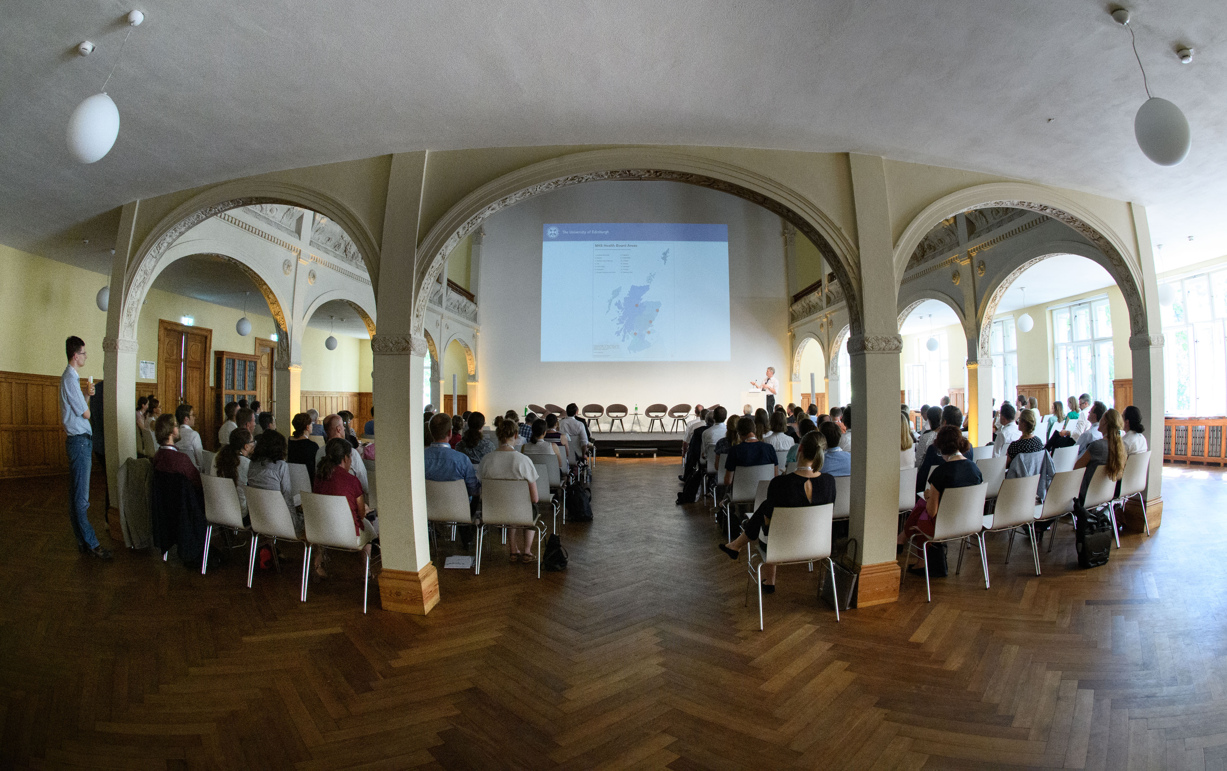 Ein Saal, in dem ein Publikum einem Vortrag lauscht