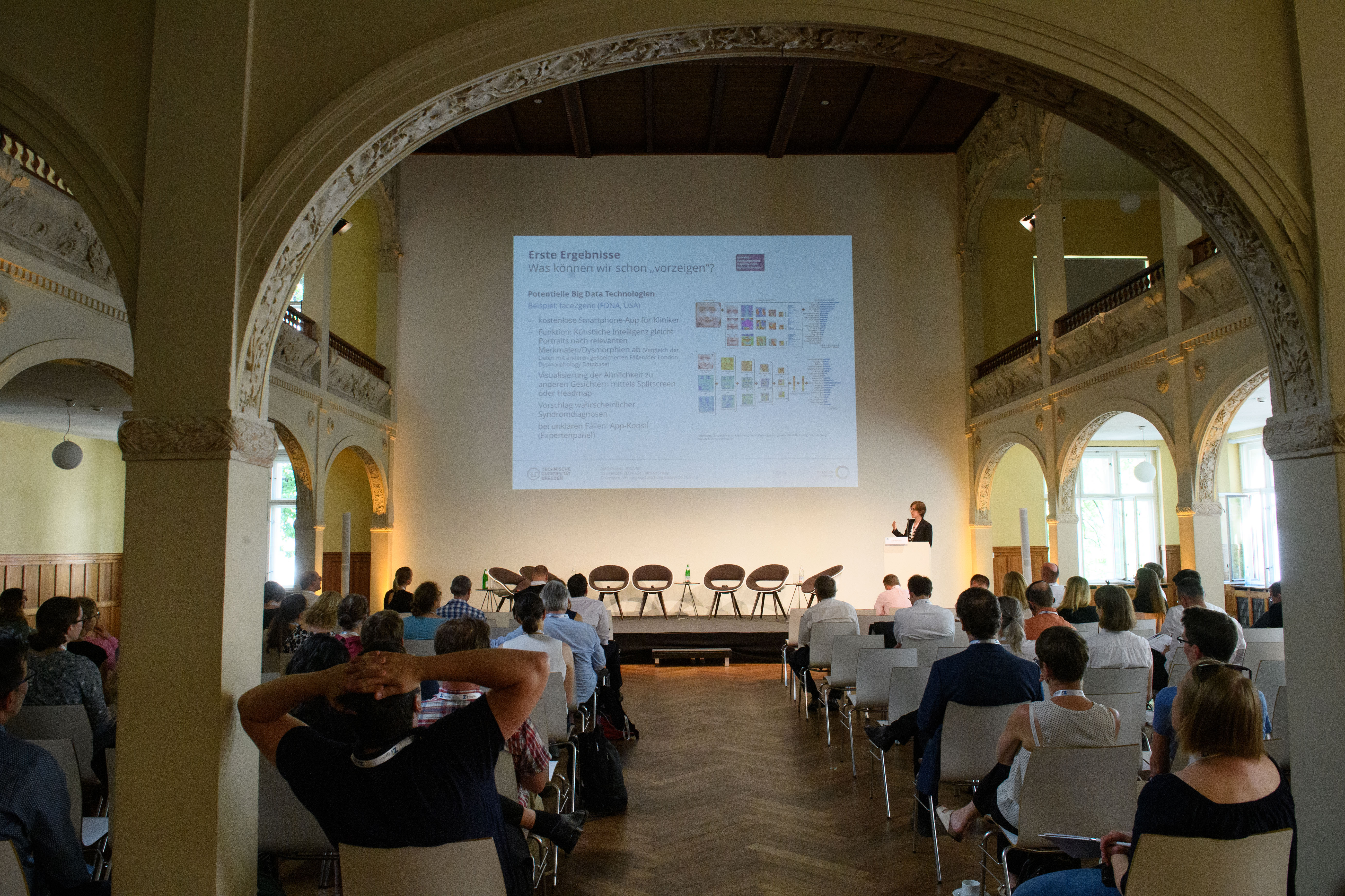 Ein Saal mit Zuhörenden, einer Vortragenden und einer Powerpointprojektion an der Wand