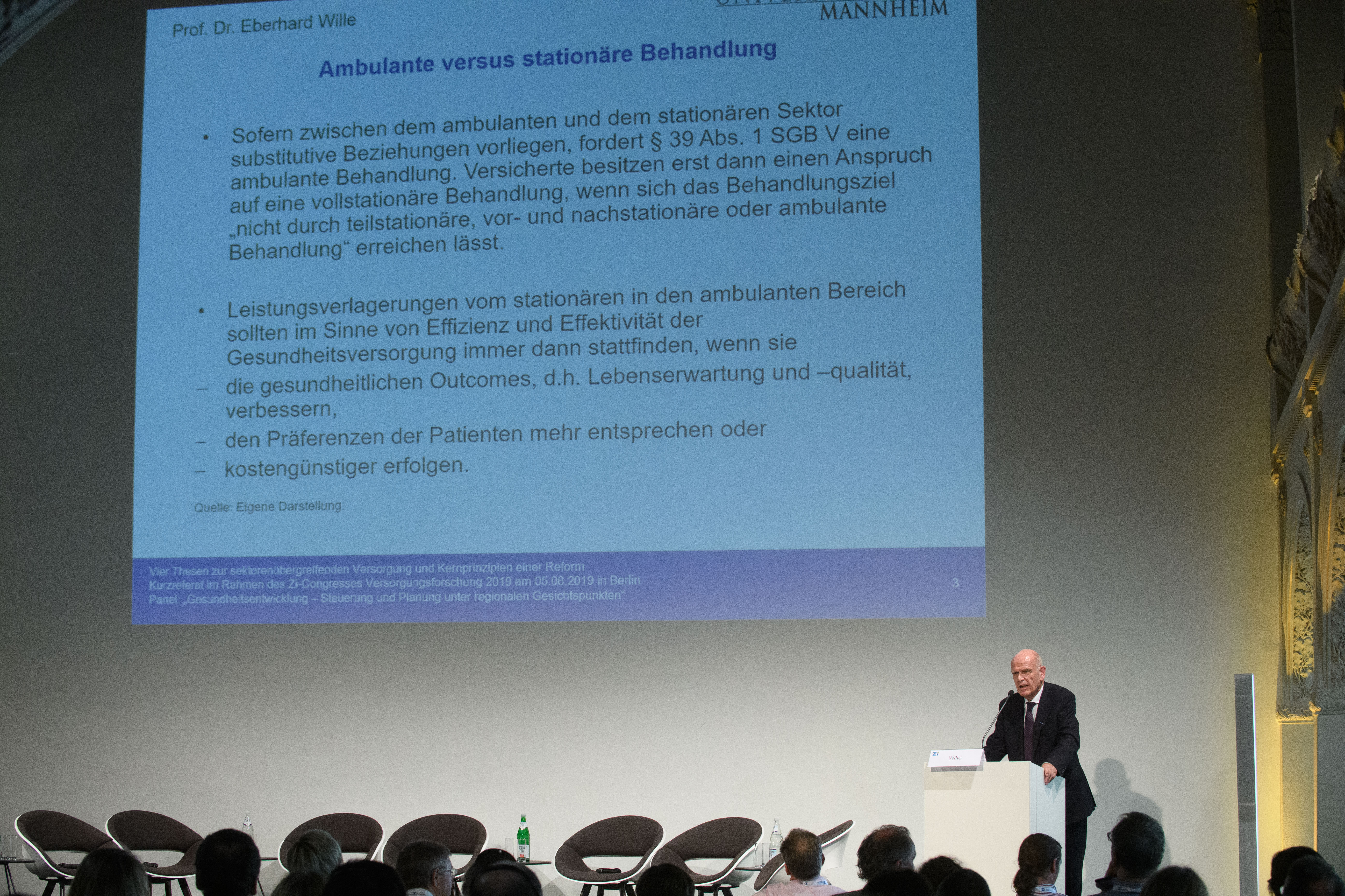 Herr Wille spricht vor einem Publikum während eine Powerpointpräsentation an die Wand projiziert wird.